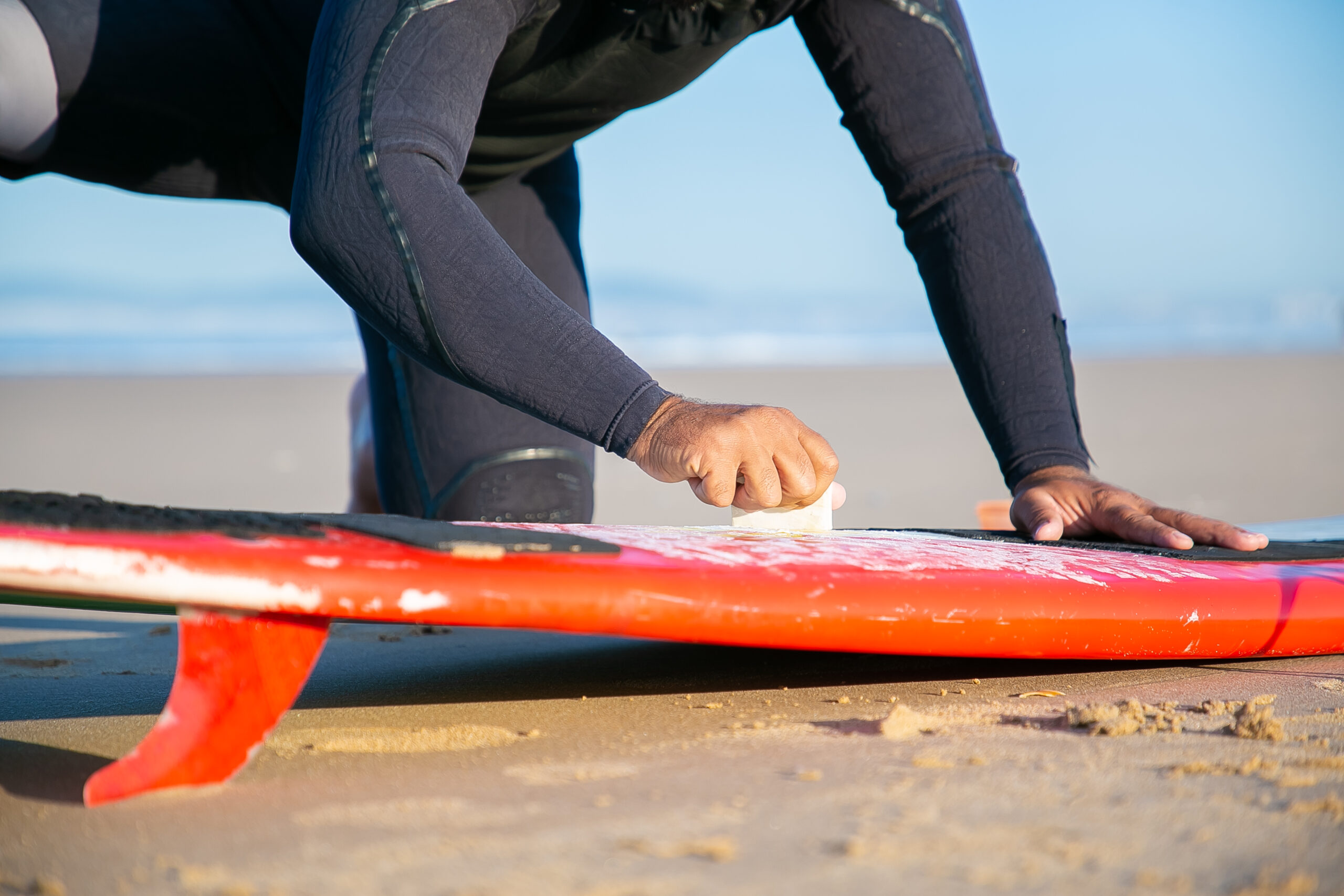 Male surfer in wetsuit waxing surfboard on sand on ocean beach.
